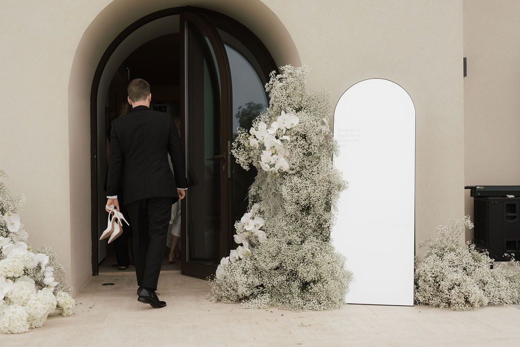 Bräutigam trägt die Schuhe der Braut und betritt die Location vorbei an einer Blumeninstallation