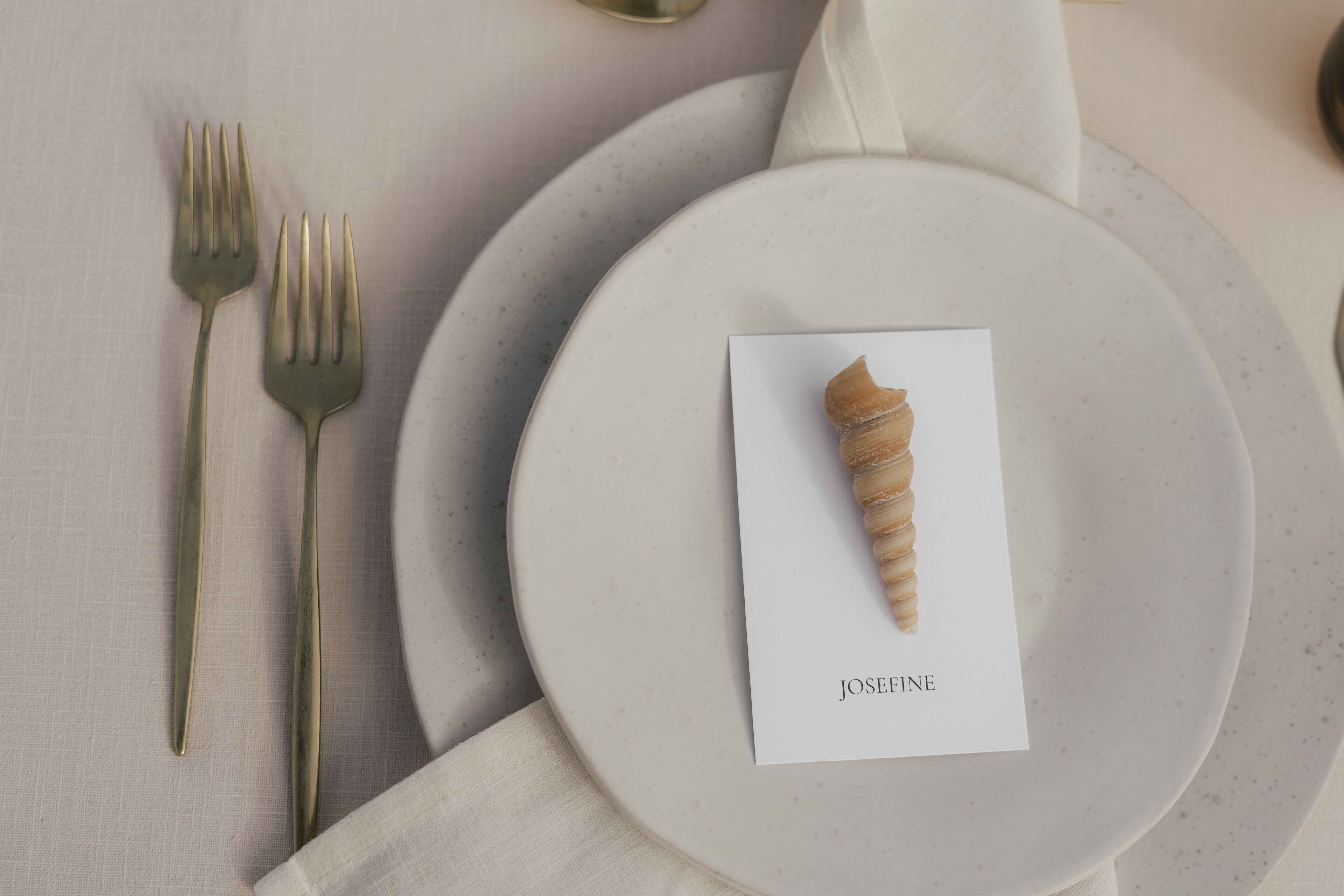 Platzkarte mit dem Namen Josefine und Spiralmuschel auf Keramikteller mit Messinggabeln — Hochzeit von Nina und Julian